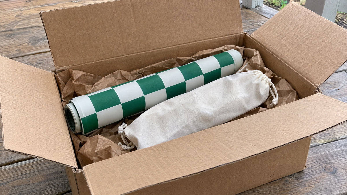 An opened shipping box on a porch holding a rolled chess board and a drawstring bag of pieces